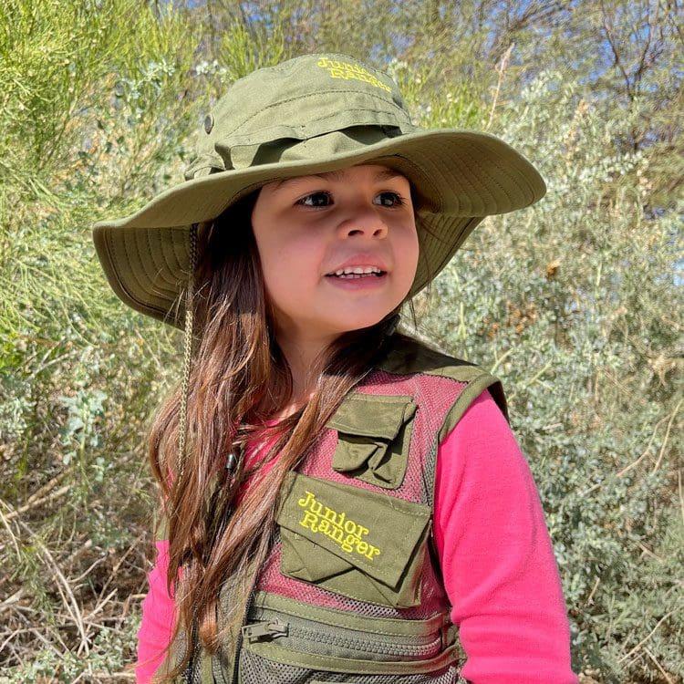 Young girl in a green hat and vest, smiling outdoors among greenery.