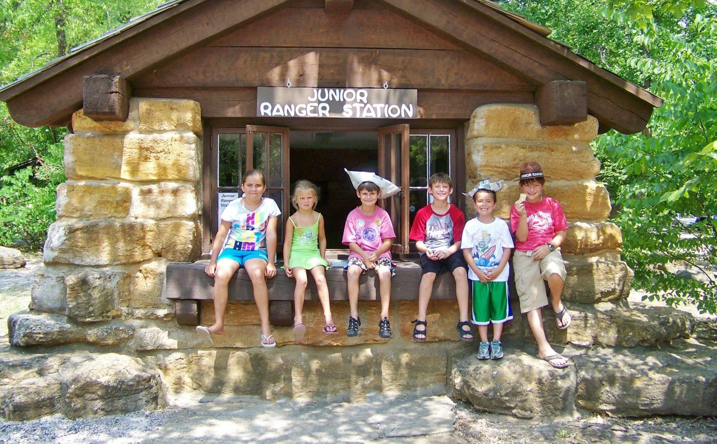 Kids sitting on a wooden cabin porch, surrounded by trees in daylight.