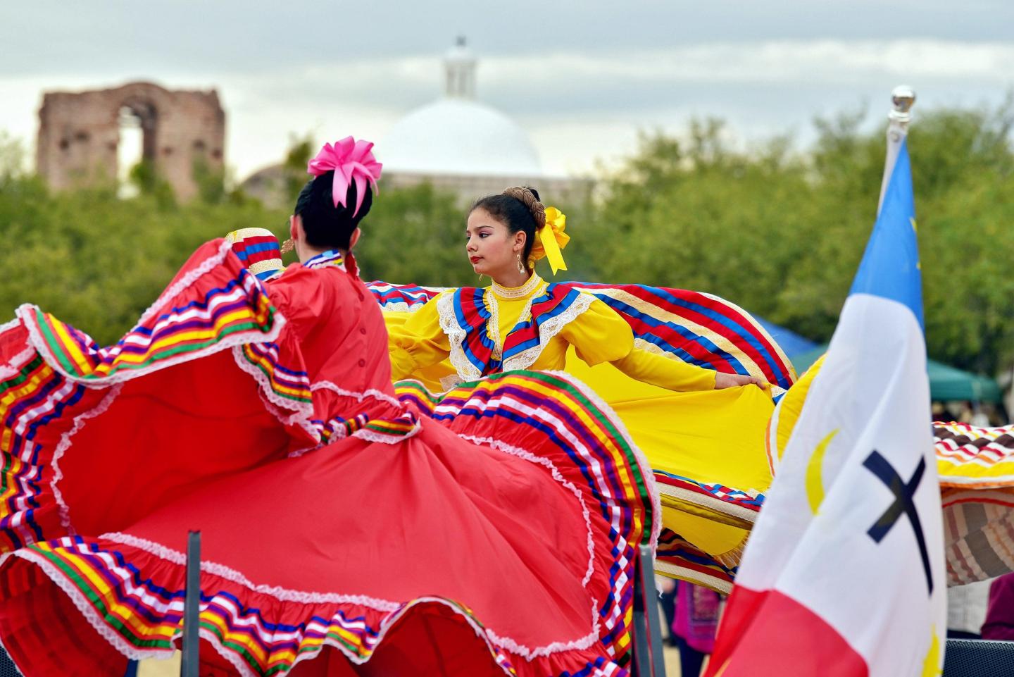 Dancers in vibrant traditional dresses twirl near a flag, with greenery and a dome in the background.
