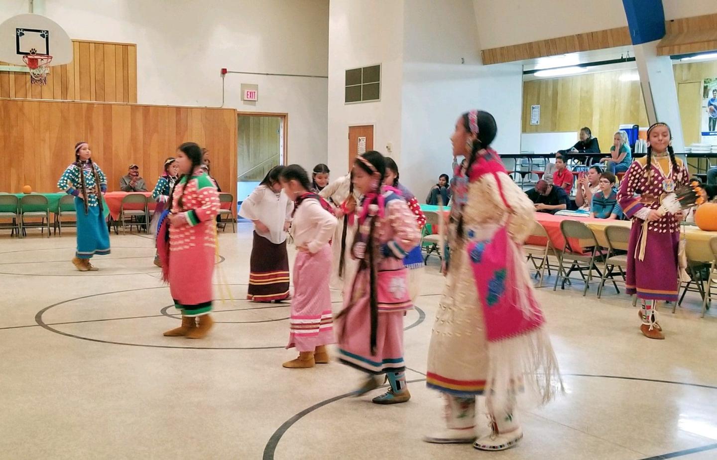 Children in traditional attire dancing in a gym with an audience watching.