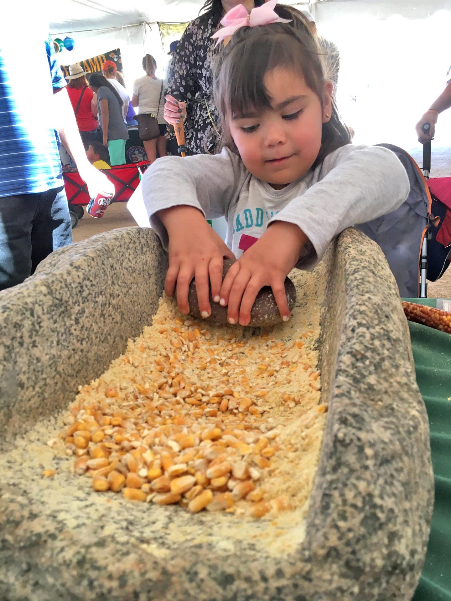 Young girl grinding corn on a stone metate at an outdoor event.
