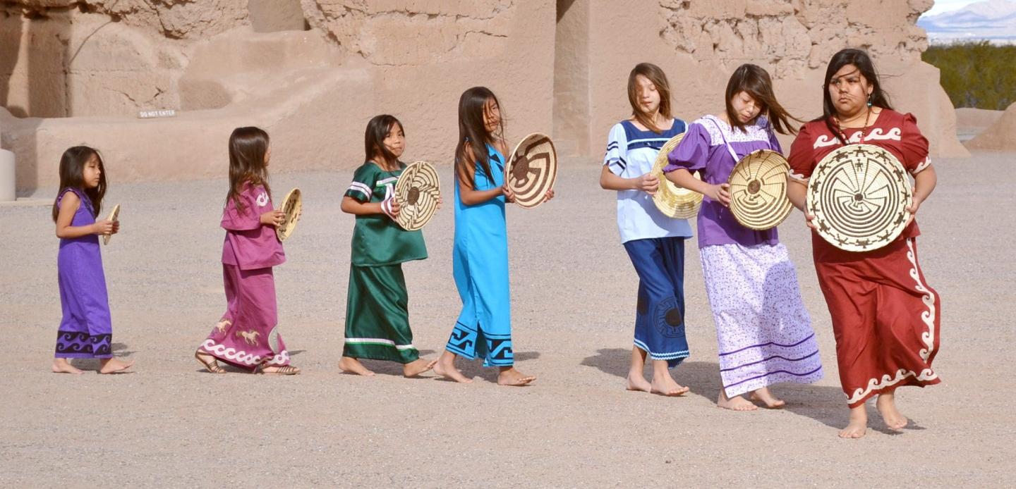 Children in colorful dresses walk in line holding drums and baskets in a desert setting.