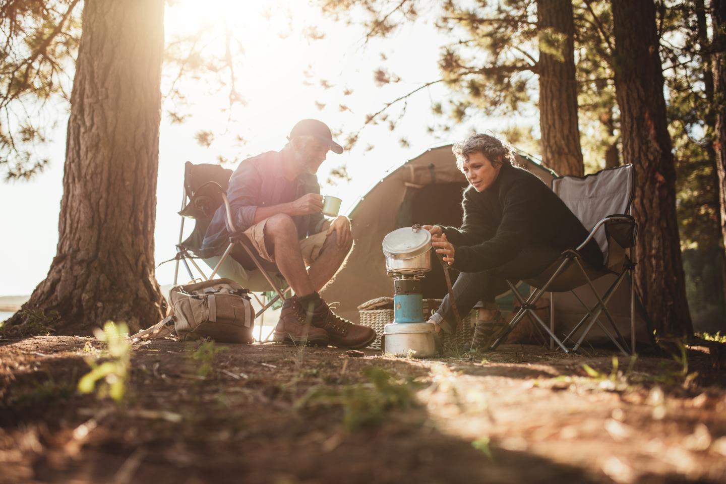 Two people camping, sitting by a tent, cooking under trees.