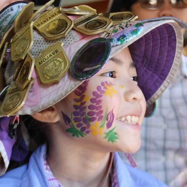 Child smiling, wearing a hat with pins, face painted with purple flowers.