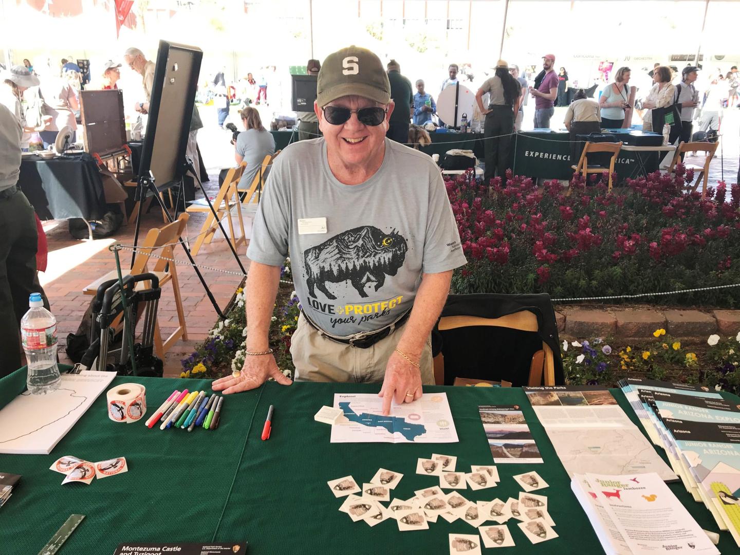 Man in cap smiling at table with brochures and stamps, outdoor event.
