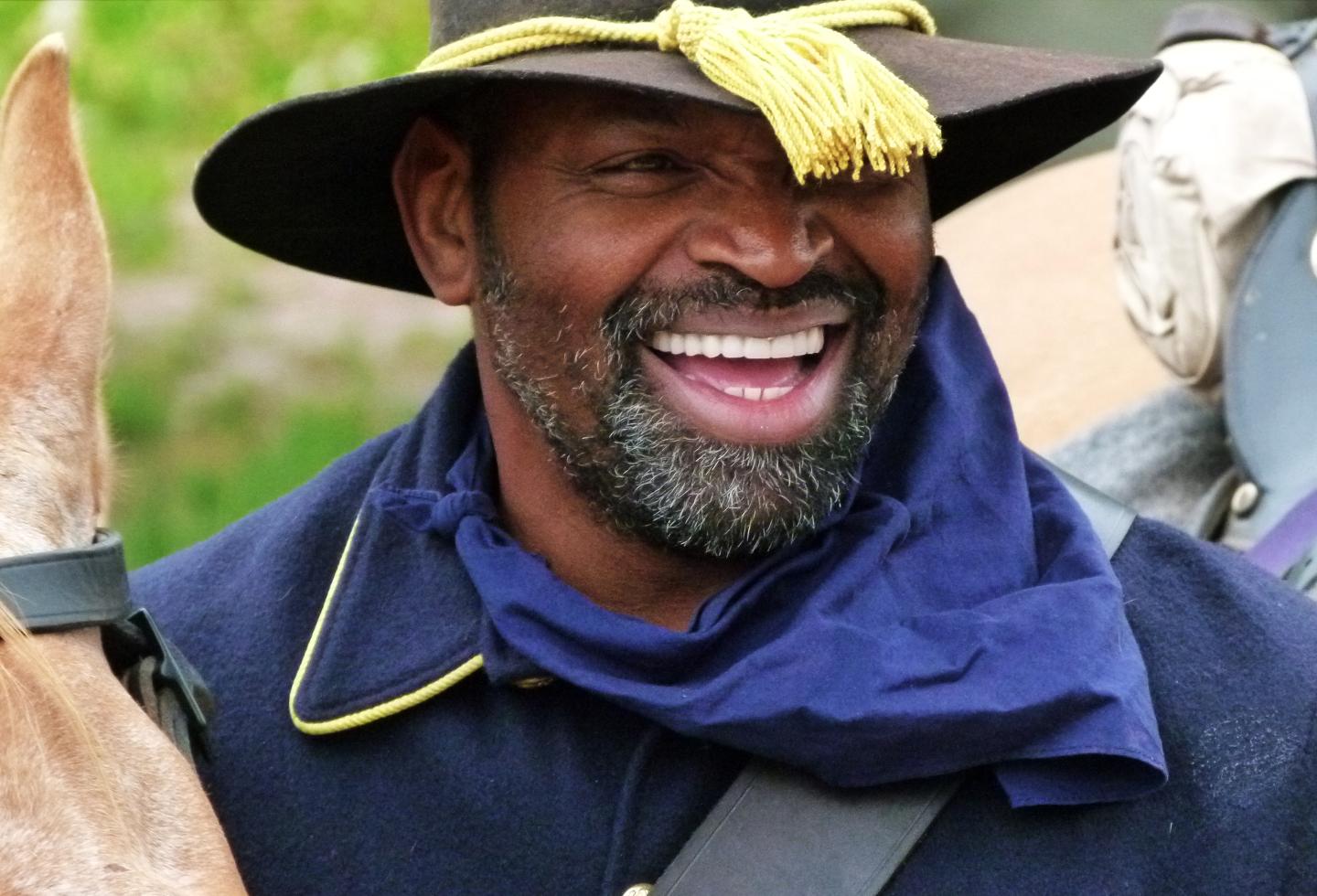 Smiling man in a blue uniform and hat holding a horse in Buffalo Soldier uniform.