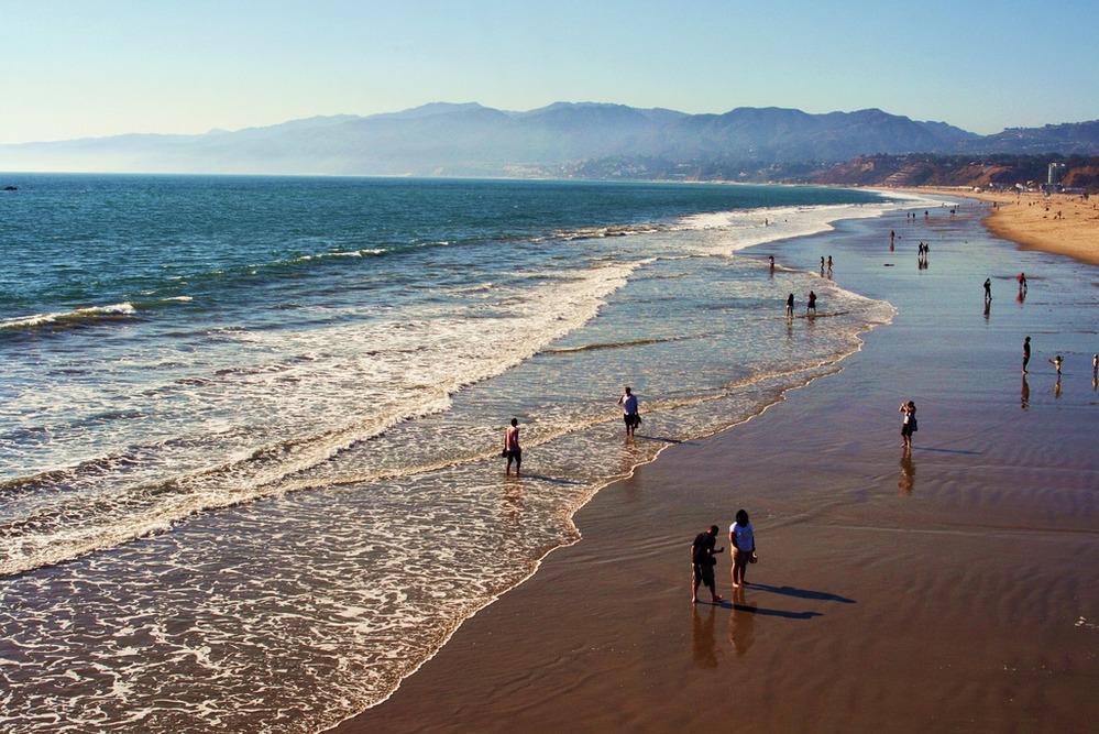 Sandy beach with people walking near gentle ocean waves. Mountains in the background.