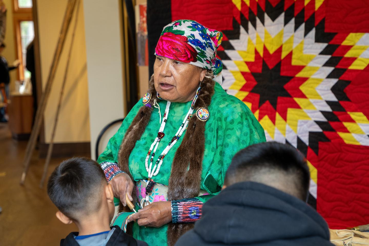 Colville Reservation woman in traditional Native American dress talks to children, colorful quilt behind her.