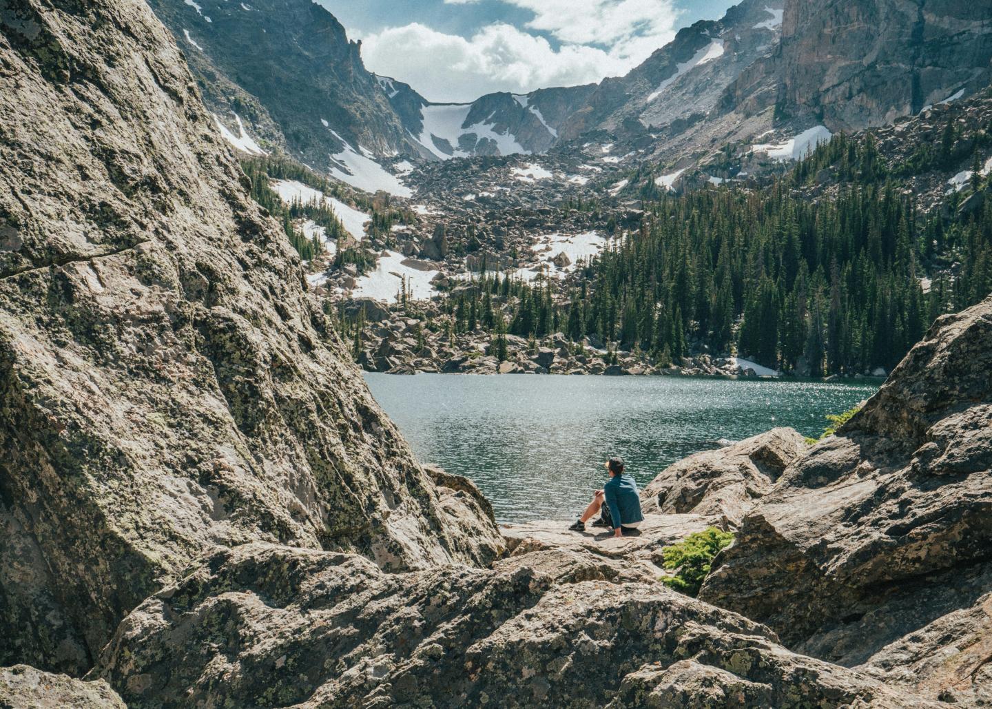 Person sits by lake, surrounded by rocky cliffs and evergreen trees.