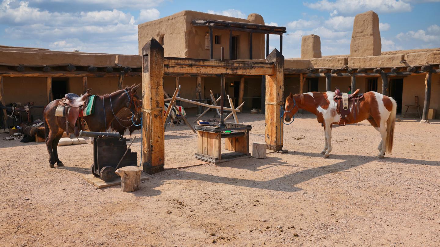Two saddled horses at Bent's Old Fort under a partly cloudy sky.