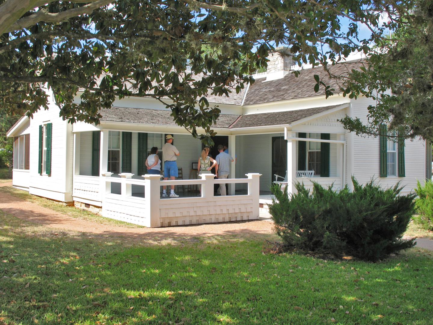 LBJ's Boyhood Home with people on porch surrounded by trees and lawn.
