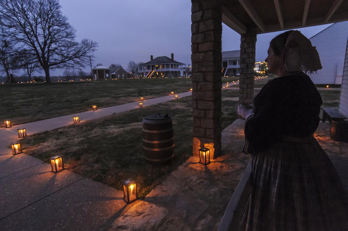 Woman in historical attire stands by candle-lit pathway at dusk.