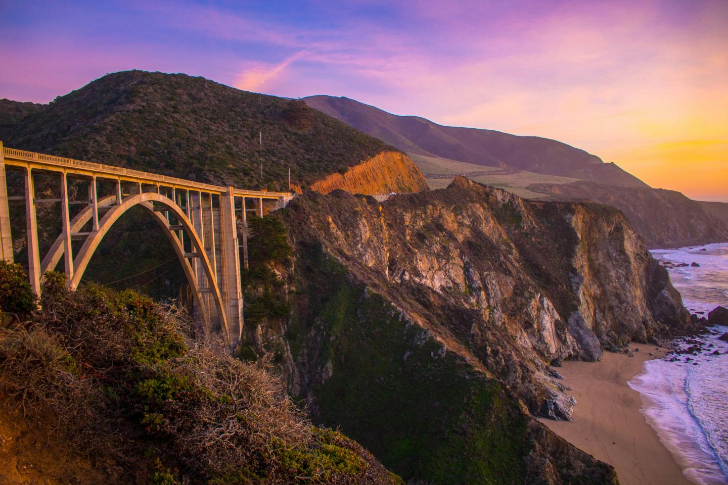Bridge over rocky coastline at sunset with purple and orange sky.