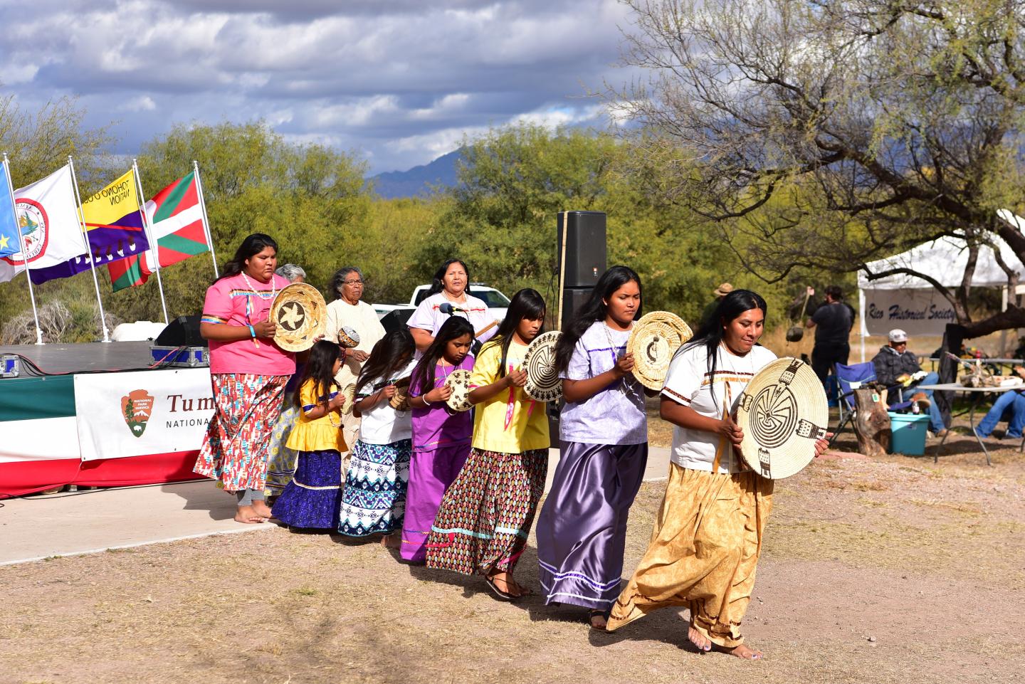 A group performs in colorful traditional attire, holding drums outdoors.