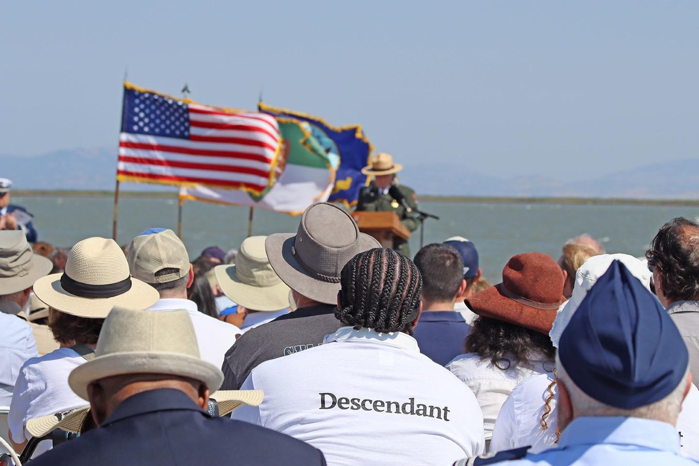 Outdoor gathering with seated crowd and speaker on stage near flags at Port Chicago Naval Magazine National Memorial