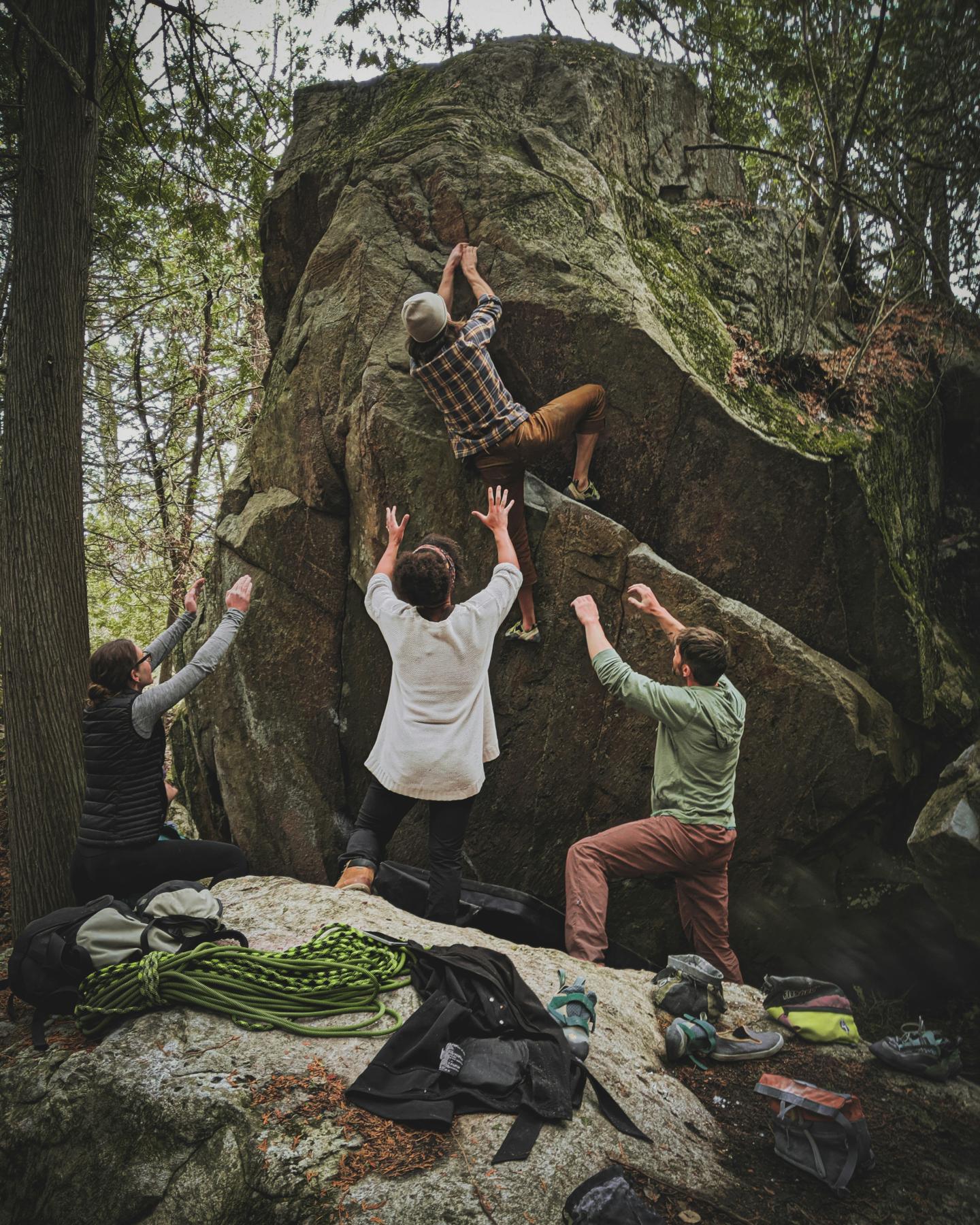 Rock climber scales a boulder with three people spotting, surrounded by trees.