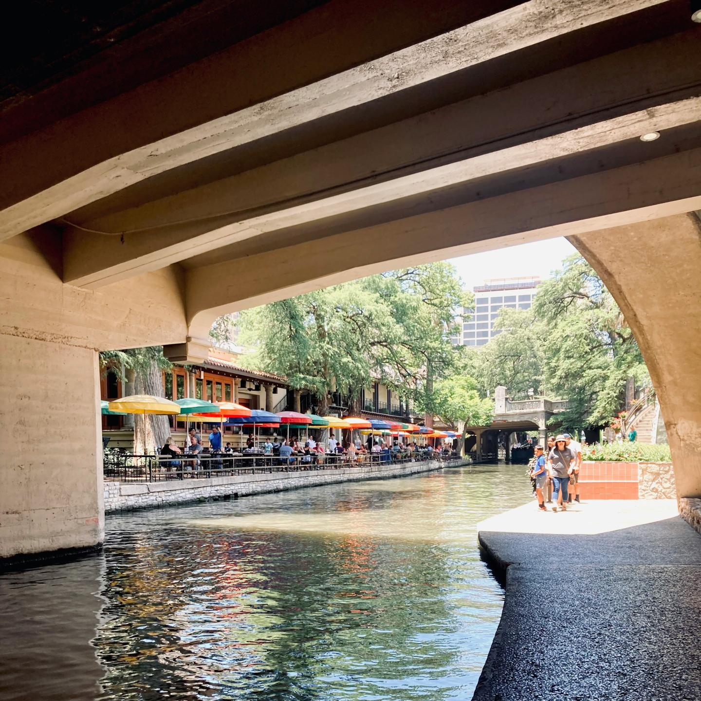 Riverwalk with colorful umbrellas by a canal walkway under a bridge.