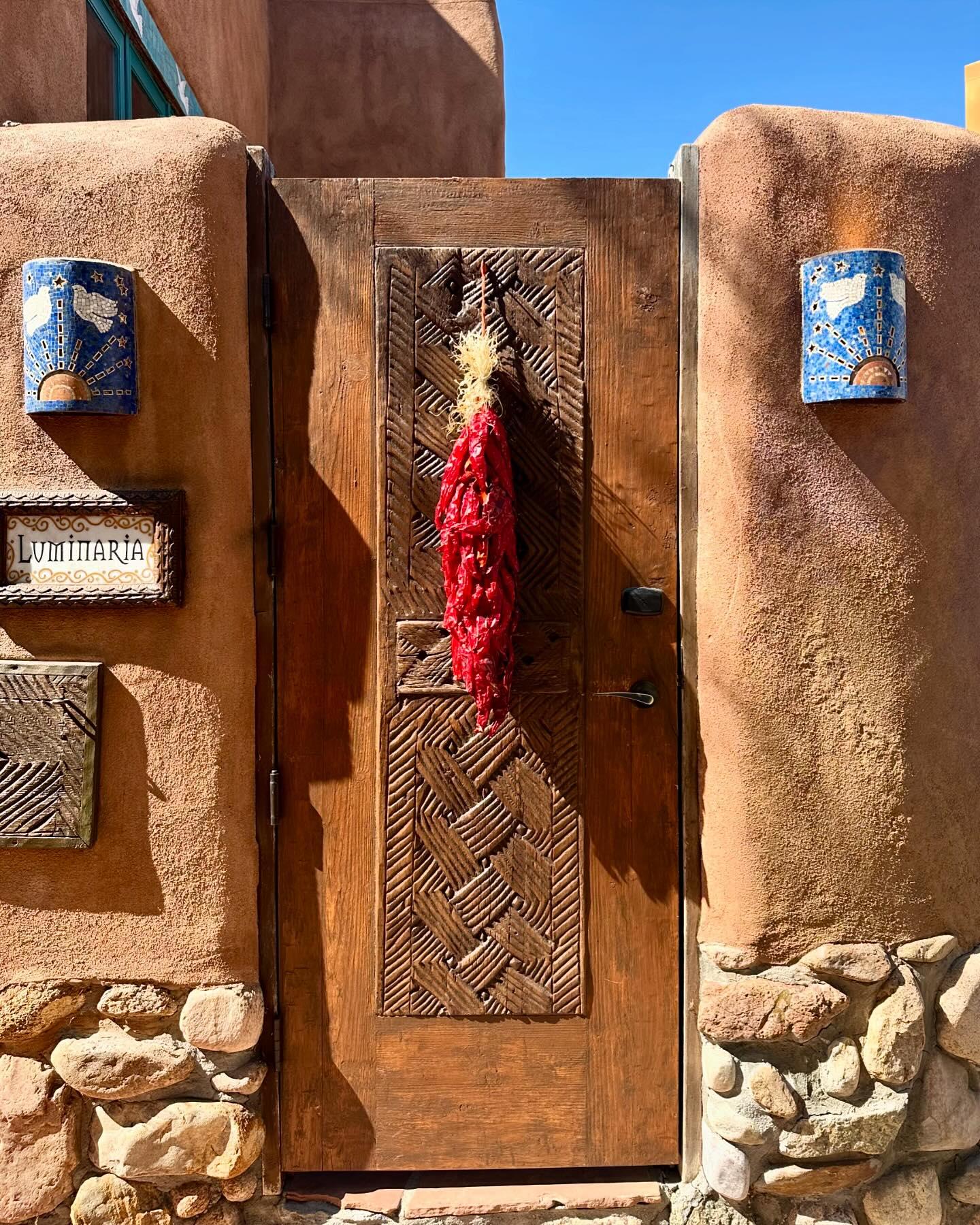 Rustic wooden door with red chili peppers hanging, adobe walls under bright blue sky.
