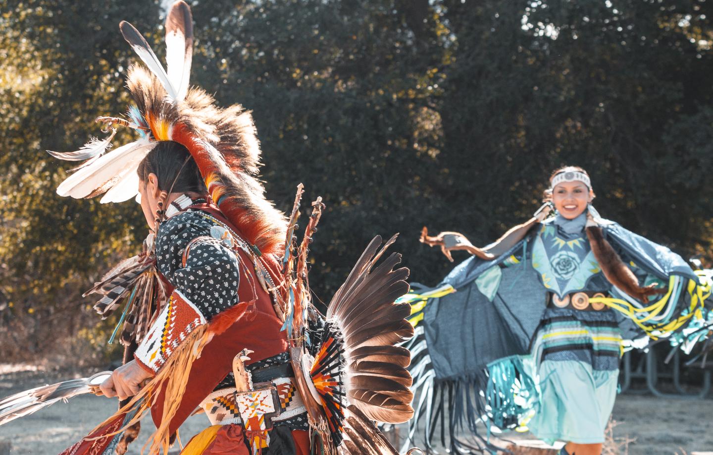 Dancers in traditional attire perform outdoors, colorful regalia with feathers and bright patterns.