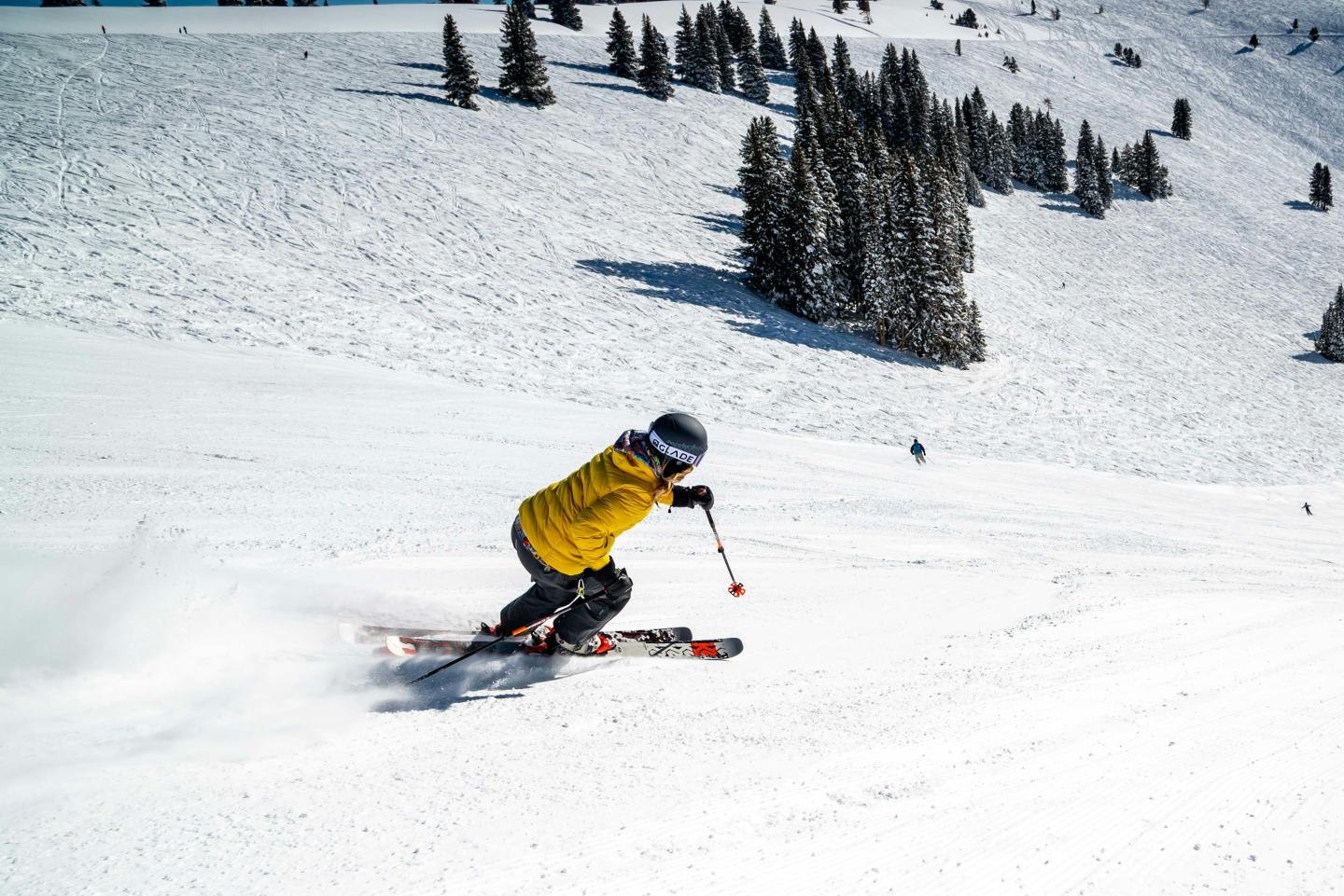 Skier in yellow jacket on snowy slope, clear sky, pine trees in background.