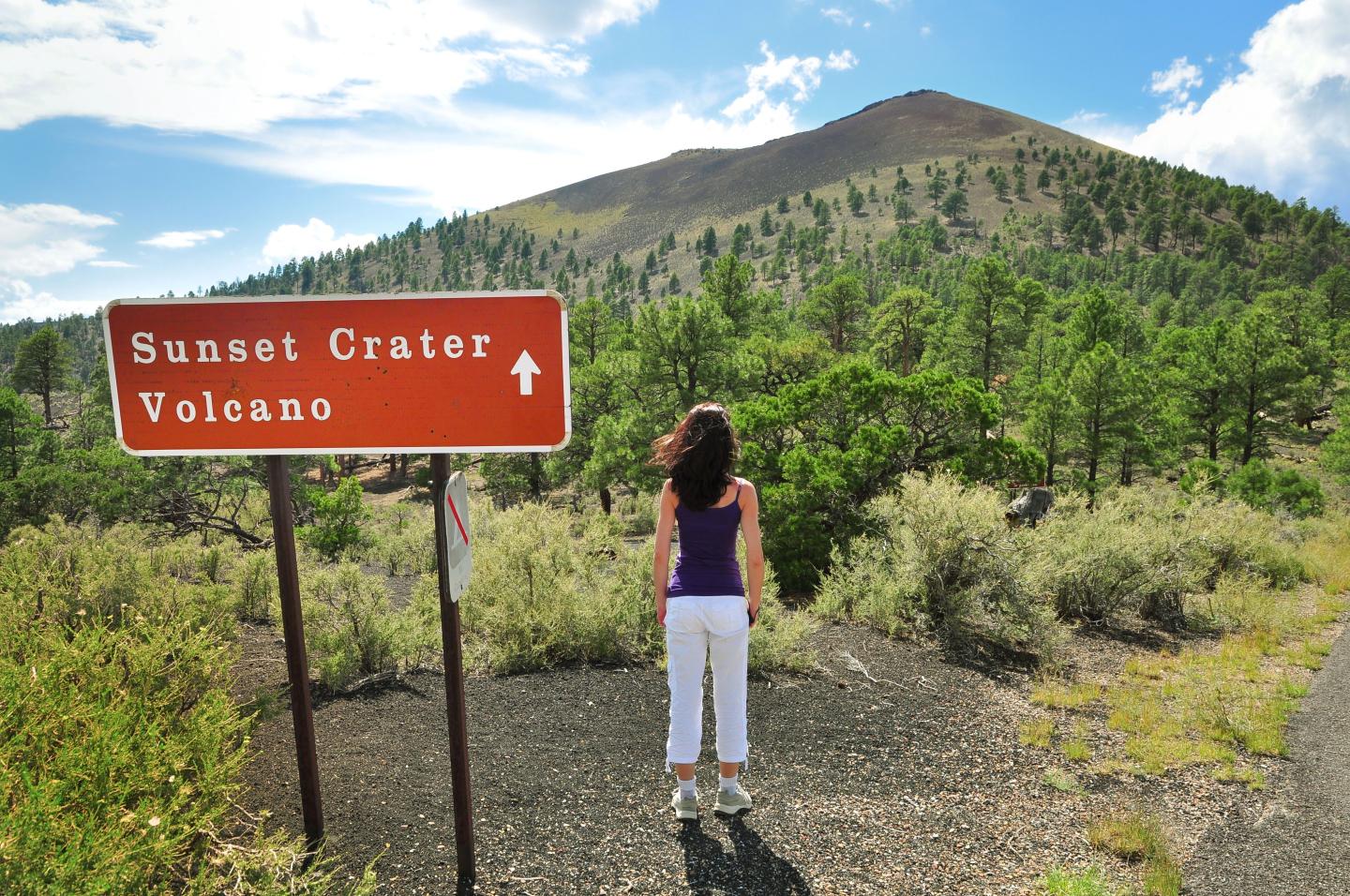 Woman standing by a sign for Sunset Crater Volcano, with a mountain in the background.