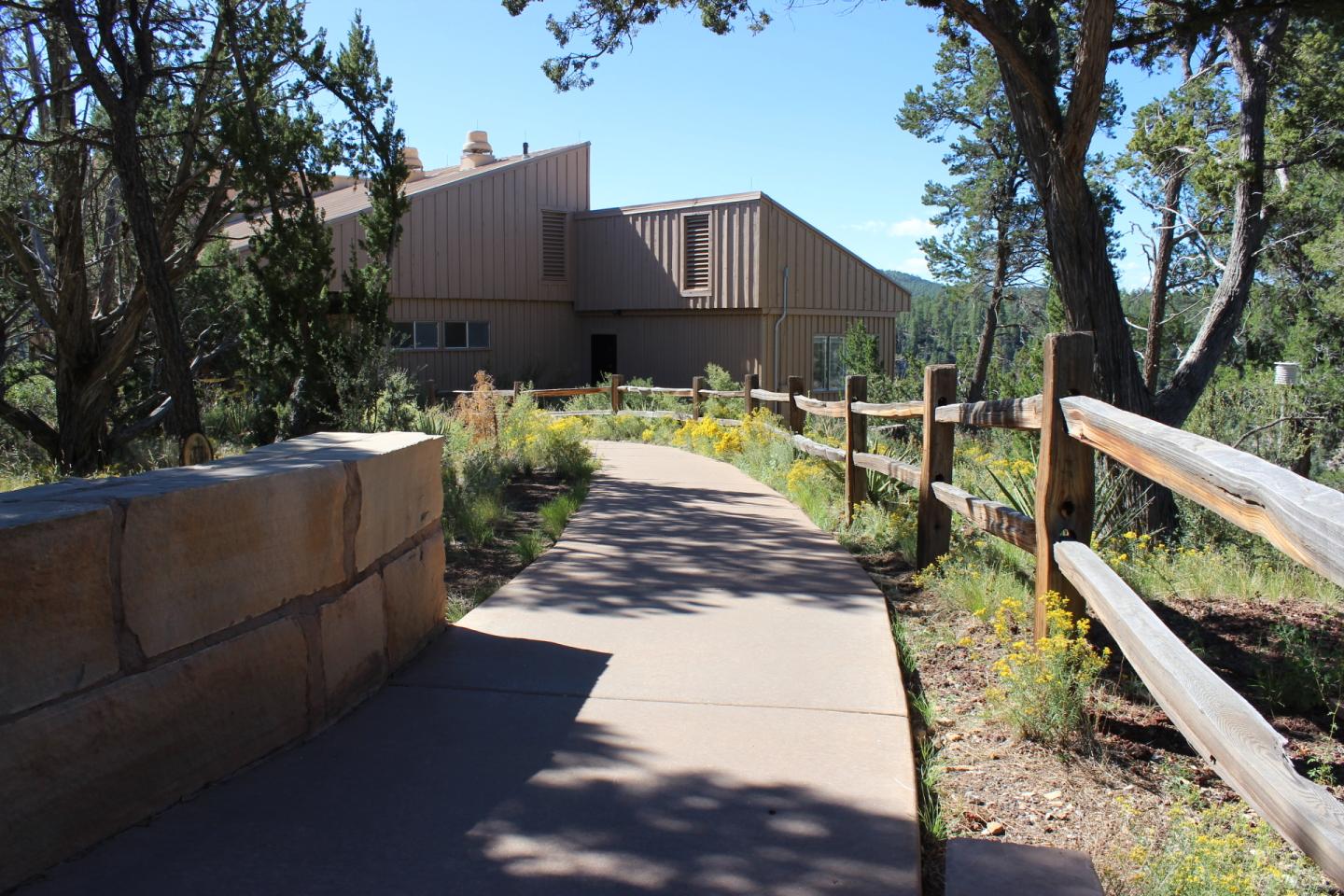 Paved path leading to a modern building, flanked by wooden fences and wildflowers.