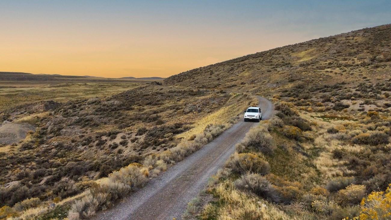 Car on dirt road through scenic desert landscape at sunset.