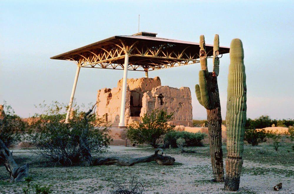 Ancient adobe ruins under a shelter with cactus and desert landscape.