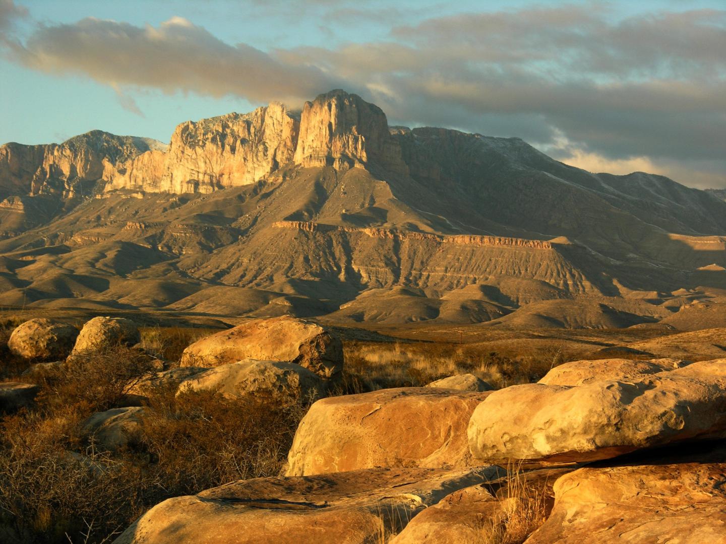 Desert landscape with rocky foreground and mountain under cloudy sky.