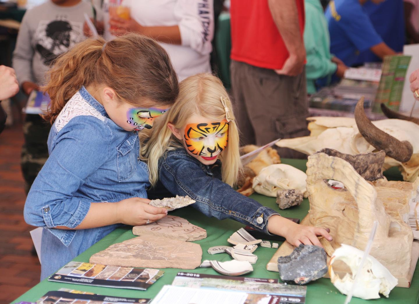 Two children with painted faces examine fossils on a table.