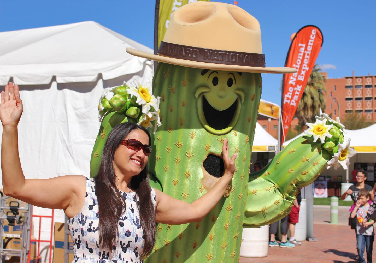 Woman posing with a smiling cactus mascot in a sombrero at an outdoor event.