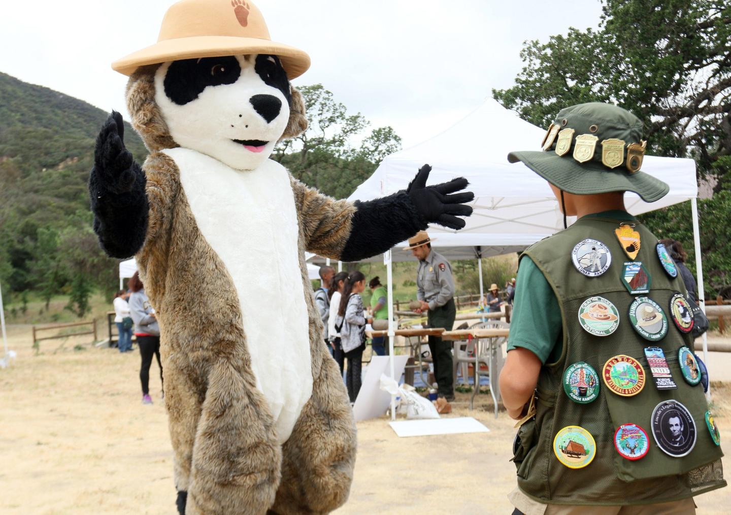 Mascot and person in badge-covered vest outdoors, with tents and trees in background.