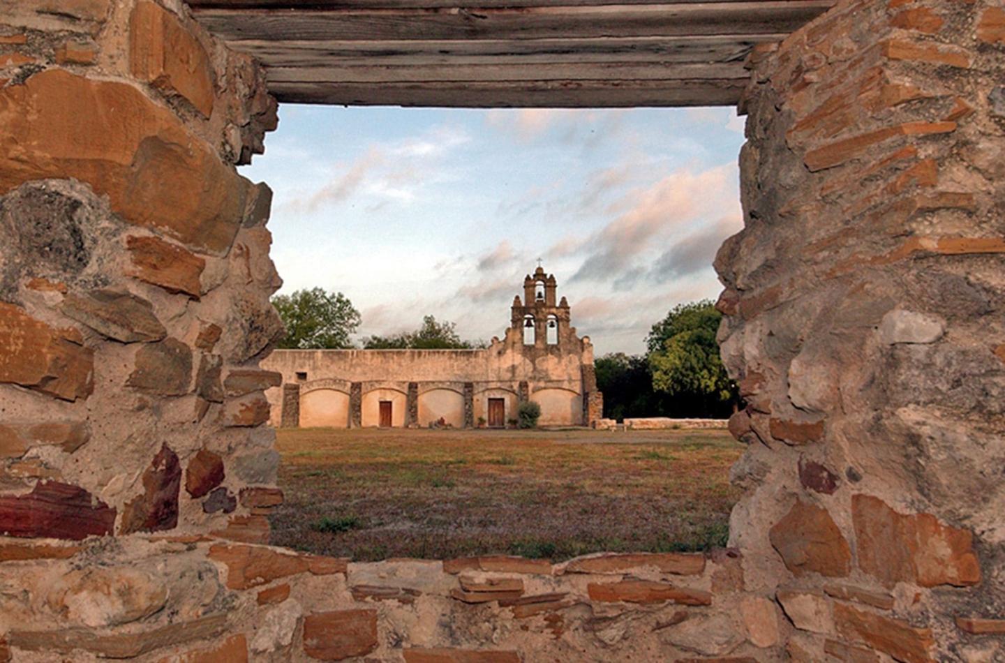 Stone ruins frame a distant historic bell tower under a cloudy sky.