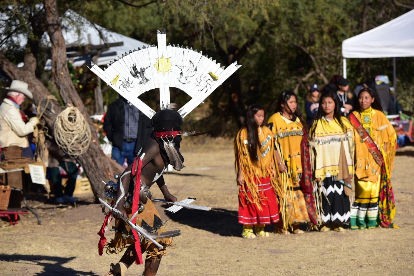 Apache crown dancer in traditional attire, with four women in colorful dresses nearby outdoors.