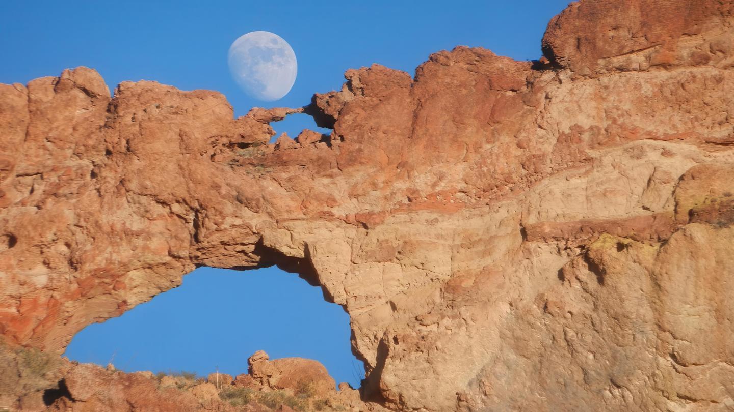 Red rock arch with moon in blue sky.