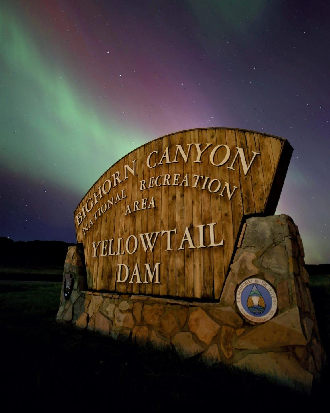 Northern lights over a wooden sign for Bighorn Canyon and Yellowtail Dam at night.