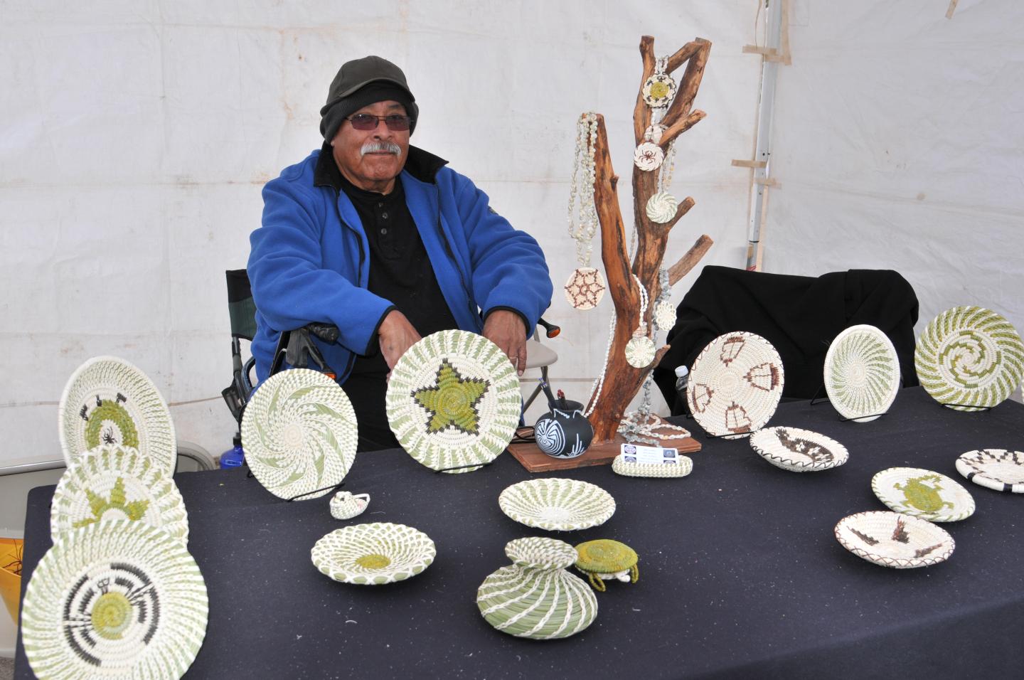 Man in a blue jacket sitting behind a display of decorative hand-woven baskets.