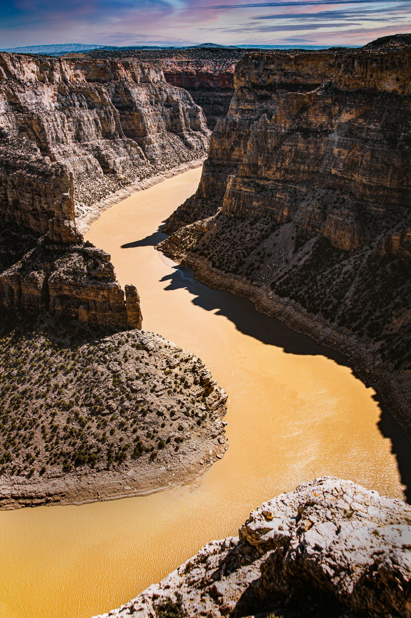 Canyon with a winding, brown river and steep, rocky cliffs under a partly cloudy sky.