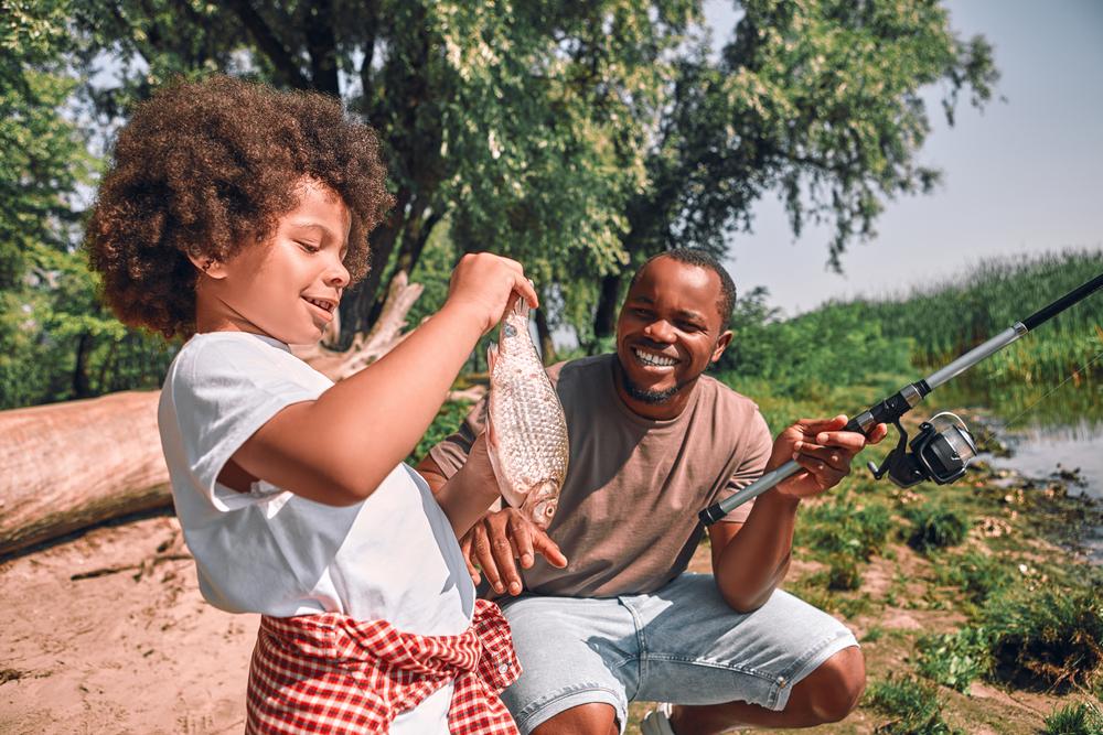Child proudly shows a fish to a smiling adult by a lakeside.