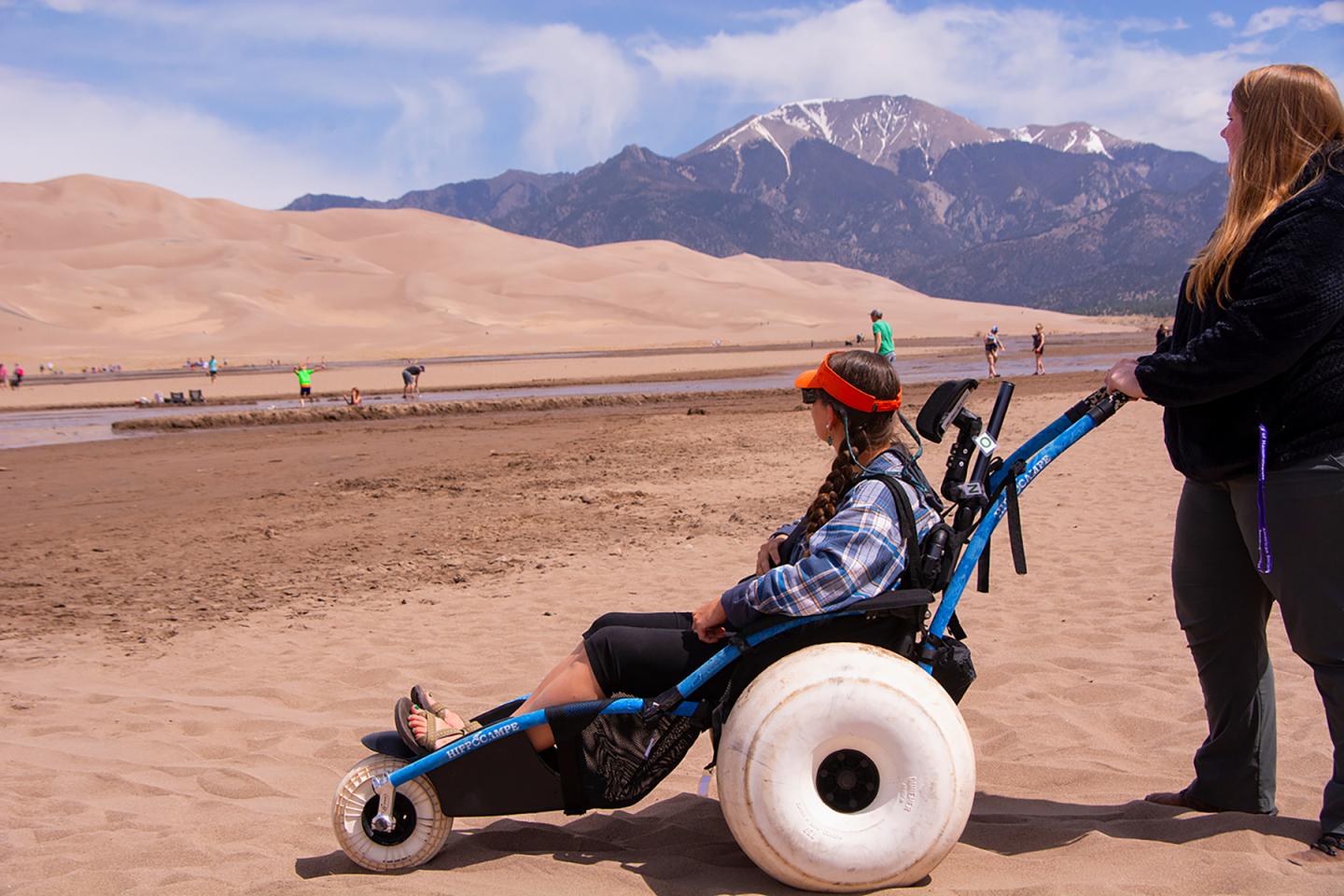 A person in a wheelchair on a sandy beach, with mountains in the background.