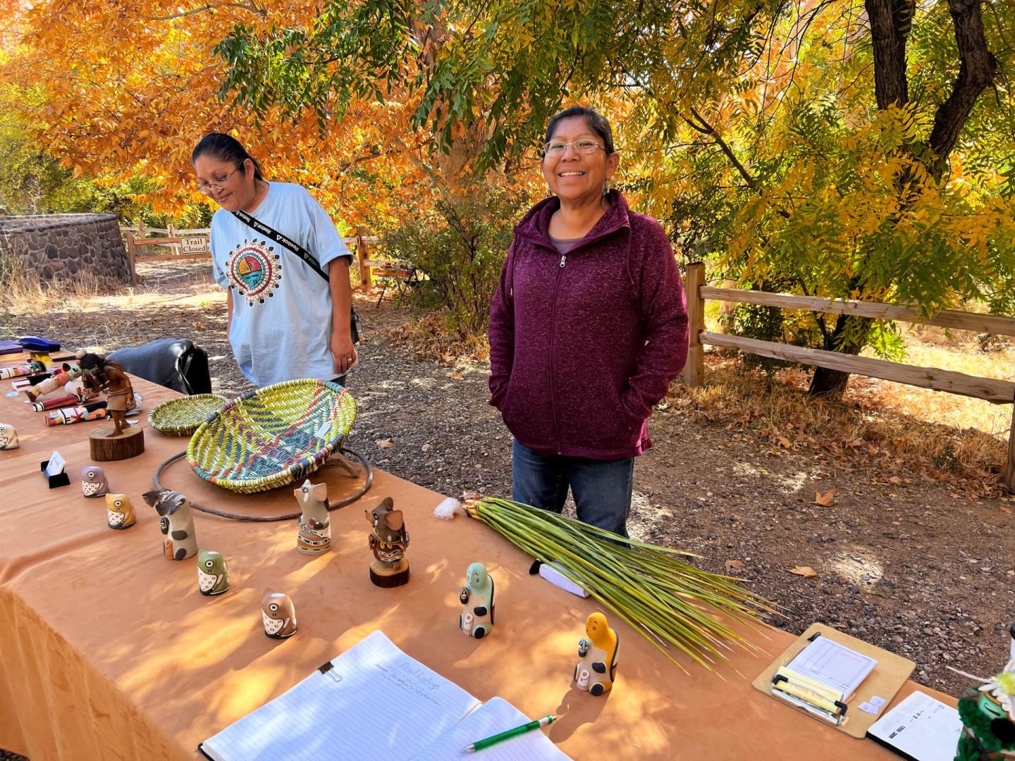 Two Native American women at an outdoor craft table with baskets and autumn trees nearby.