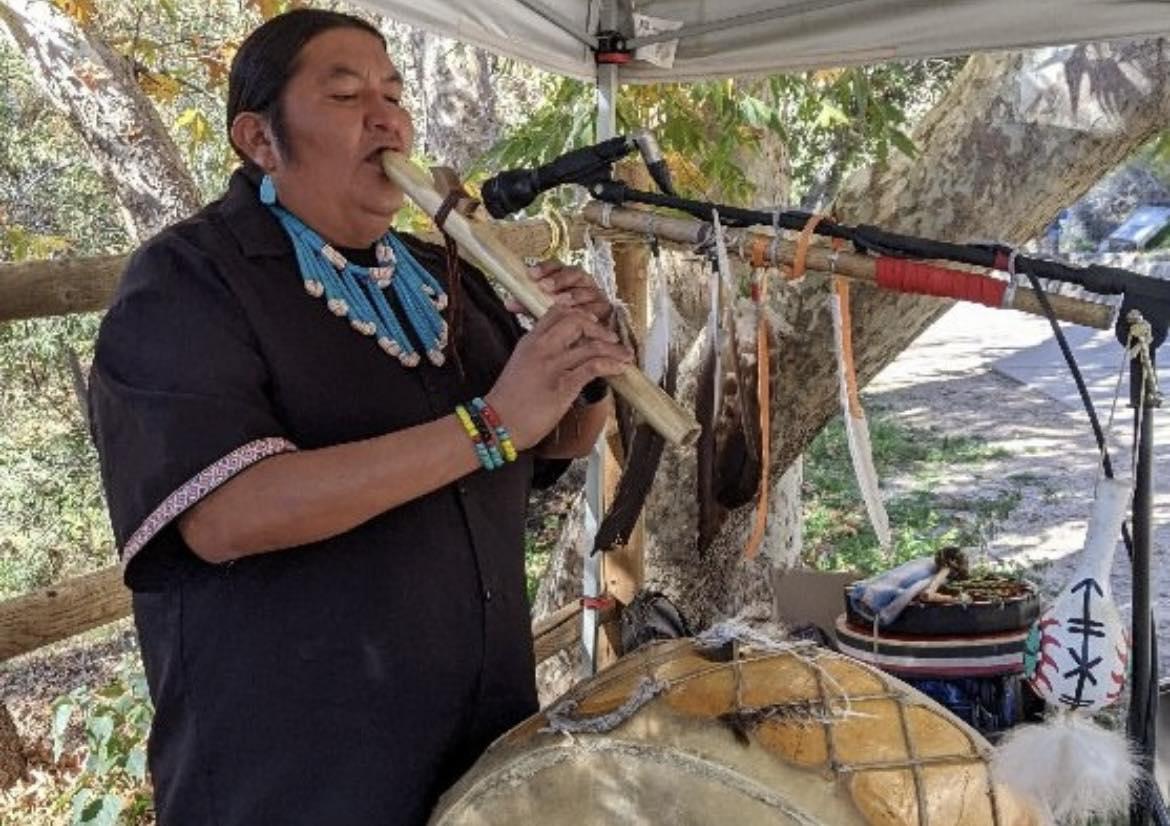 Native American musician playing a flute outdoors, surrounded by traditional items.