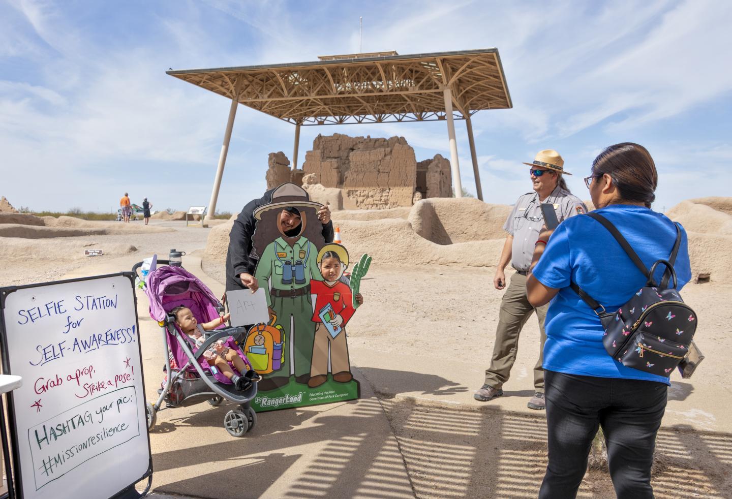 Visitors stand near a stroller and cardboard cutouts at a historic Casa Grande Ruins site under a clear sky.