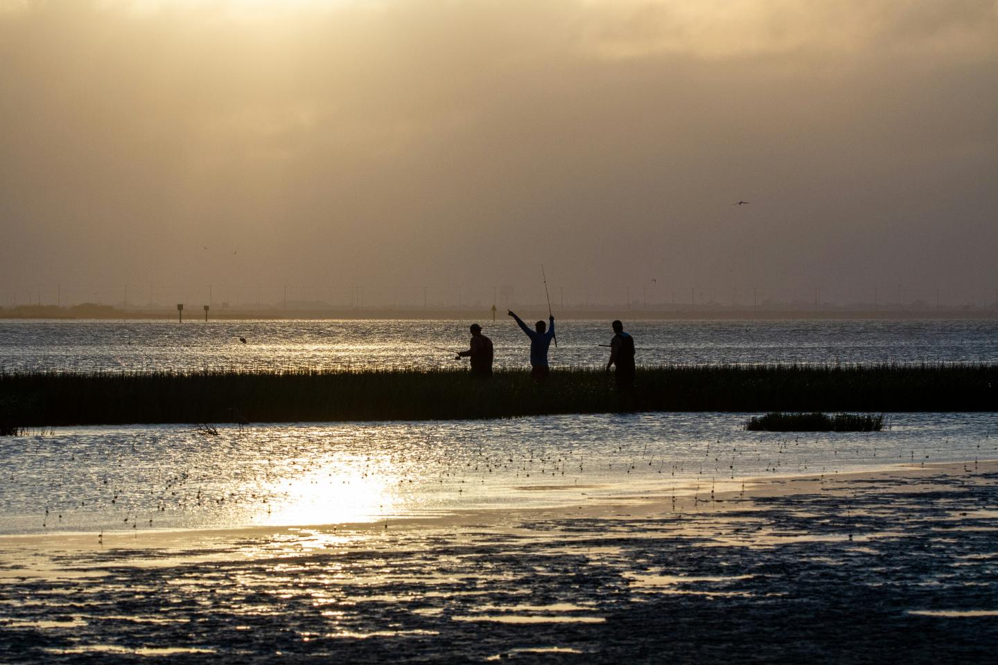 Sunset over a calm beach with silhouettes of three people on a pier.