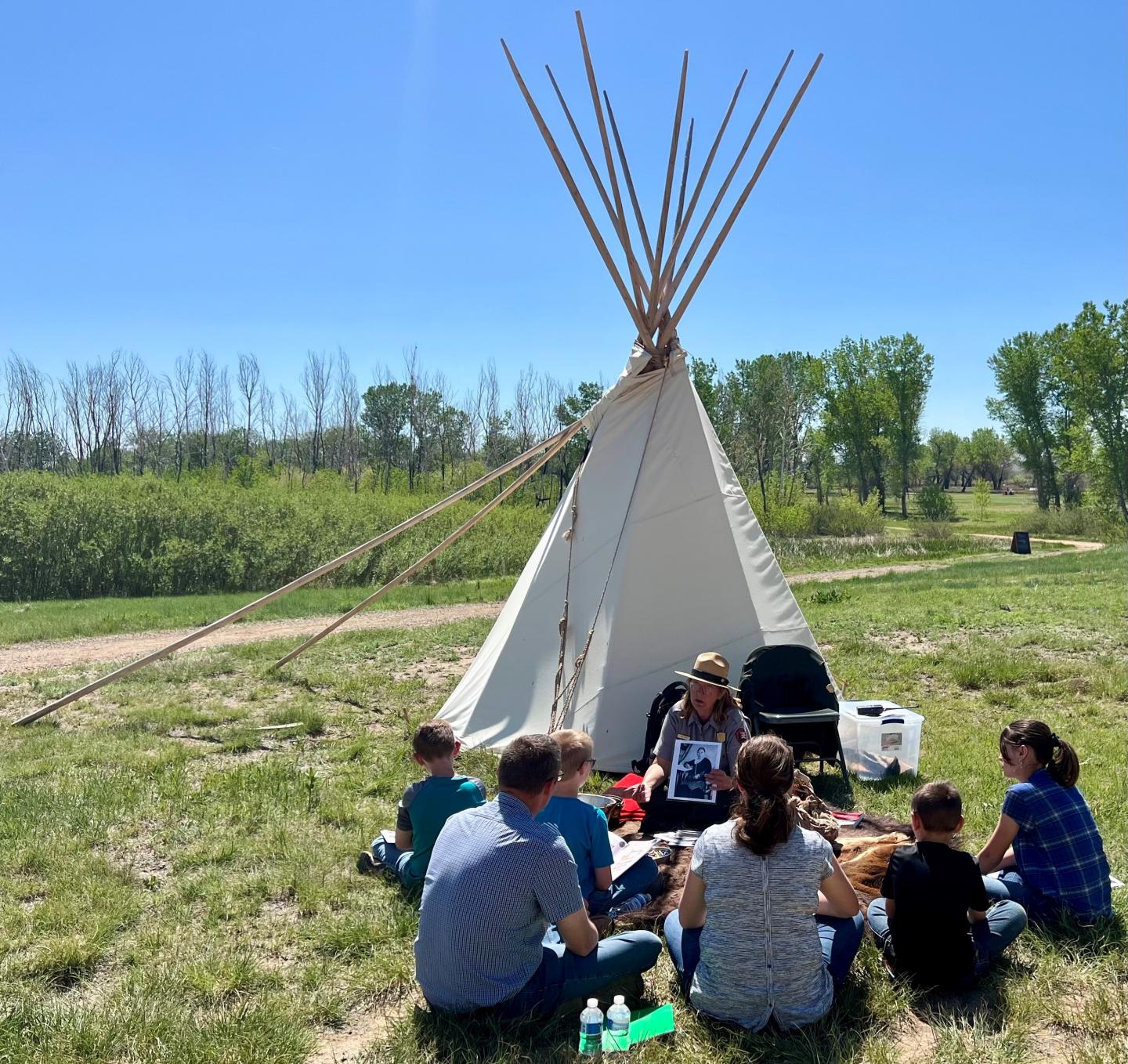 People sitting in a circle near a tipi on a sunny day in a grassy field.