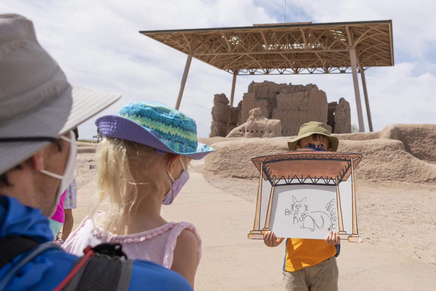 Child holding a sketch in front of ancient ruins, adults watching attentively.