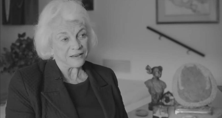 Elderly woman with white hair in a suit sits indoors, black-and-white photograph.