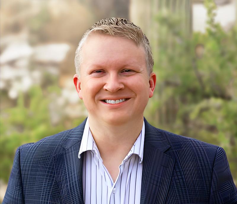Smiling person in a suit outdoors with greenery in the background.