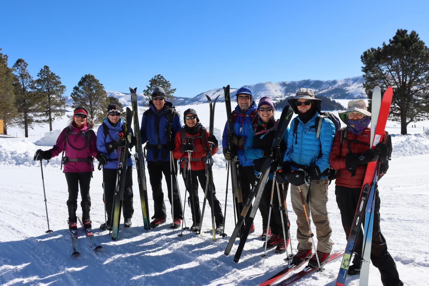 Group of skiers posing with gear on a snowy mountain under a clear blue sky.