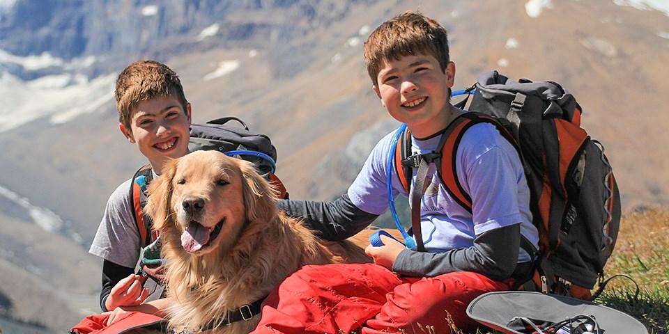 Two boys with backpacks and a golden retriever on a mountain hike, smiling.